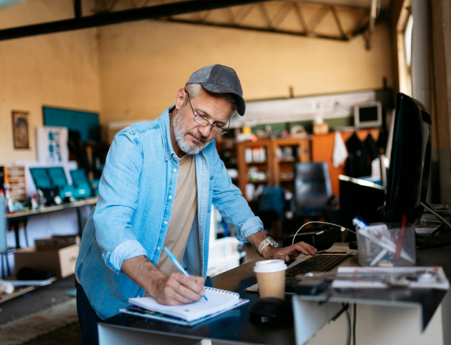 Male business owner leaning over counter, writing in a notebook in front of a coffee and computer.