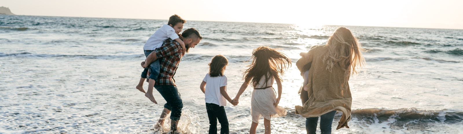 Family photo of man and woman with three young children at the beach in the water at ankle height in clothing with pants rolled up.