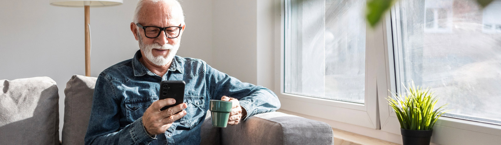 elderly man using phone sitting on couch drinking coffee